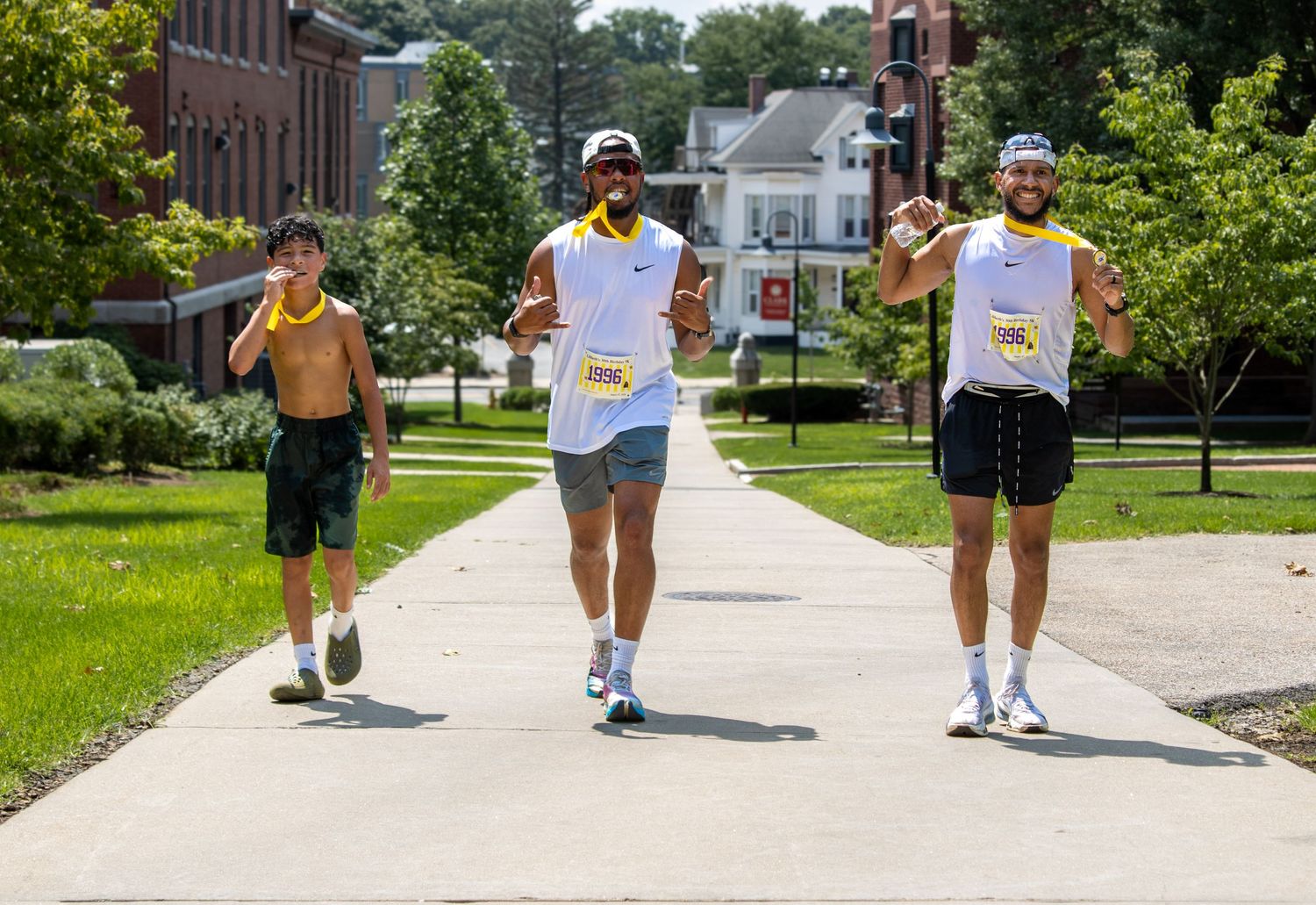 Three runners walking on a sunny campus path, two wearing race bibs, smiling and gesturing during an outdoor fun run.