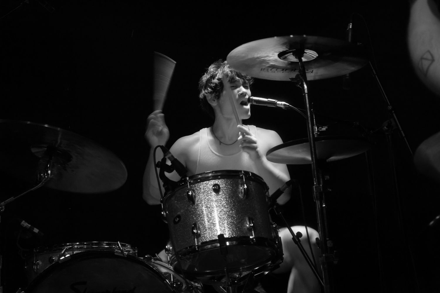 Shirtless drummer playing energetically on a sparkly drum kit on stage in black and white concert photo.