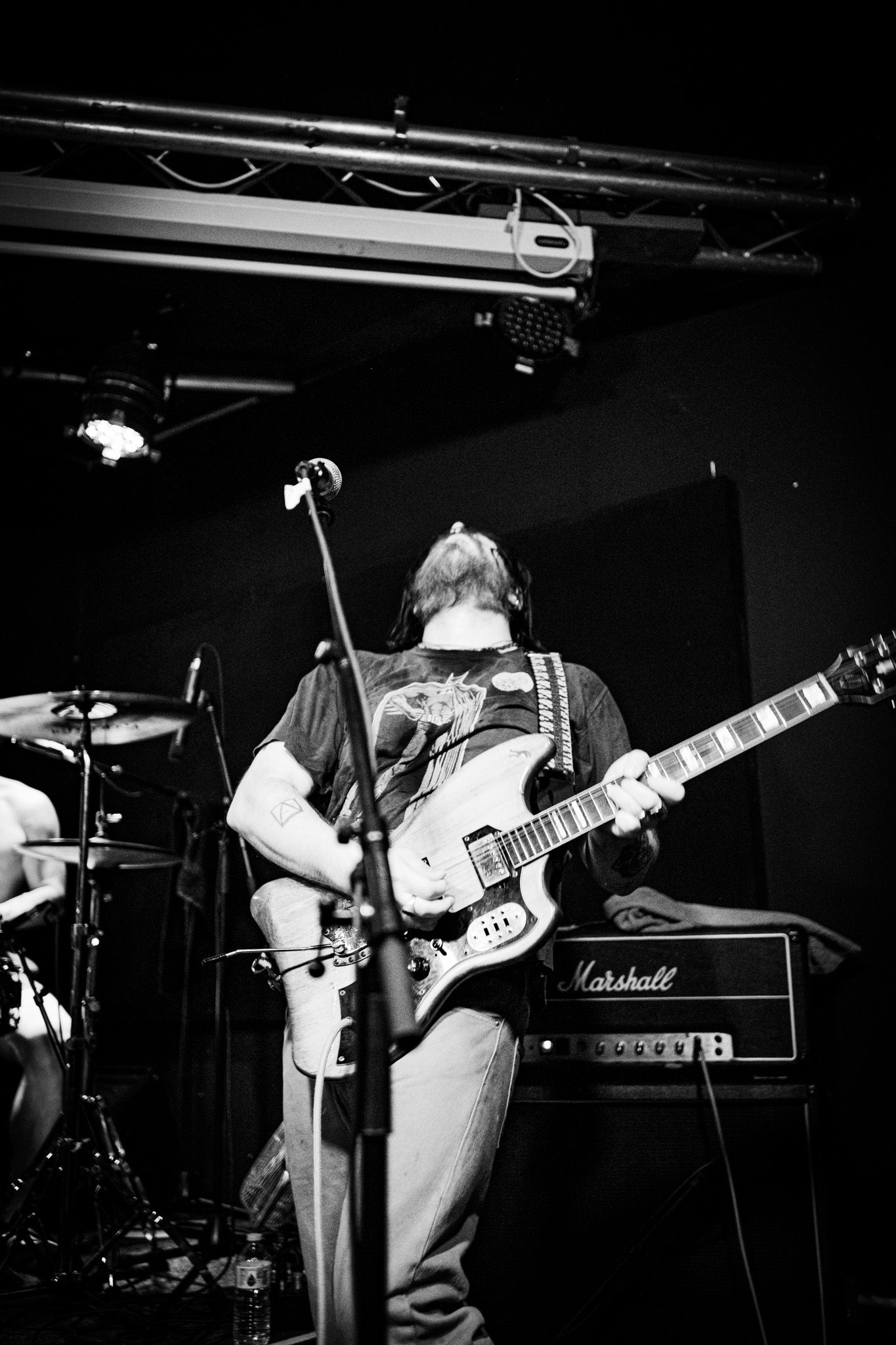 Black and white photo of a guitarist performing on stage with a Marshall amp, microphone stand, and drummer visible in background.