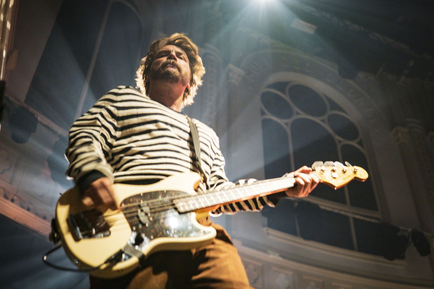 Guitarist in striped shirt performs on stage under dramatic lighting in an ornate venue.