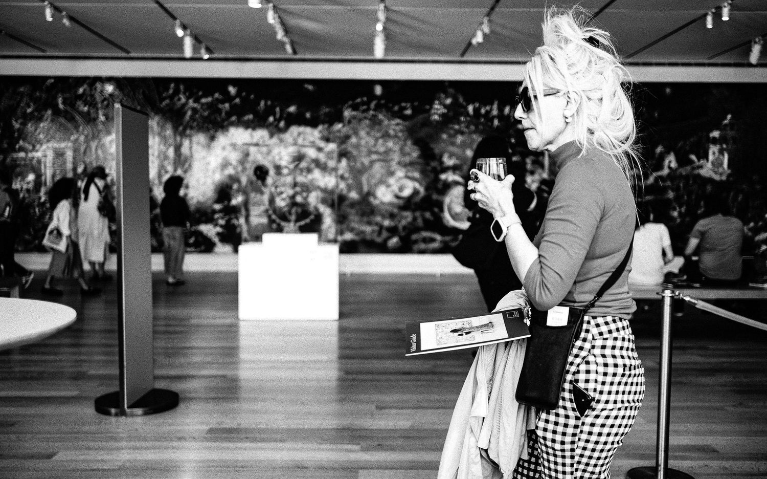 Black and white photo of blonde woman holding camera at art museum gallery exhibition with paintings in background.