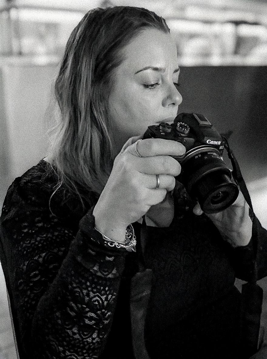 Black and white photo of a woman holding a Canon camera up to her face, wearing a black lace top.