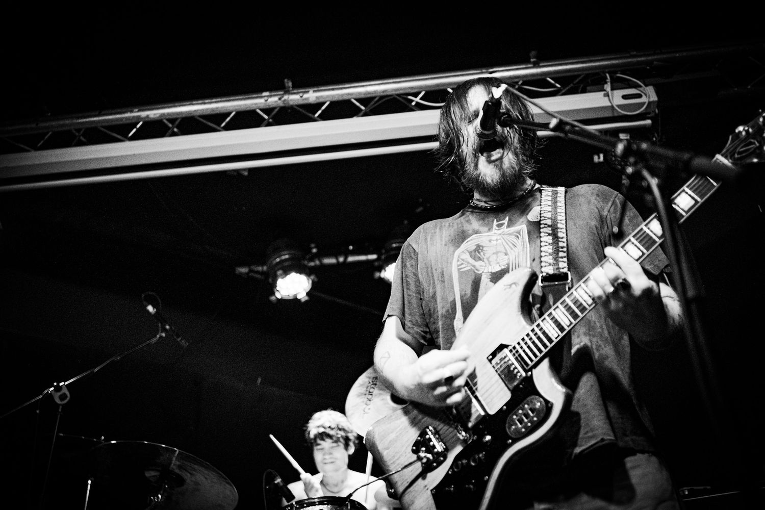 Black and white photo of a guitarist performing on stage with long hair, playing an electric guitar under bright stage lights.