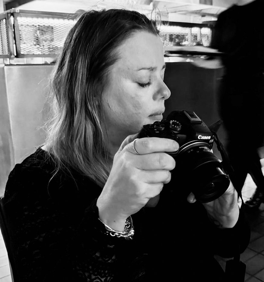 Woman in black top photographing with a Canon DSLR camera, captured in black and white.