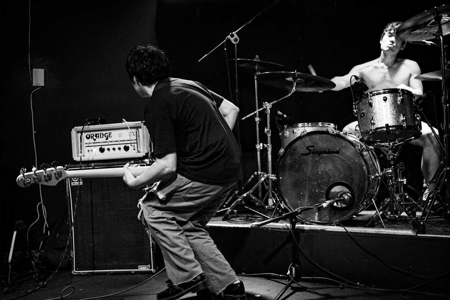Black and white photo of guitarist bending low while playing as energetic drummer performs behind kit on stage.