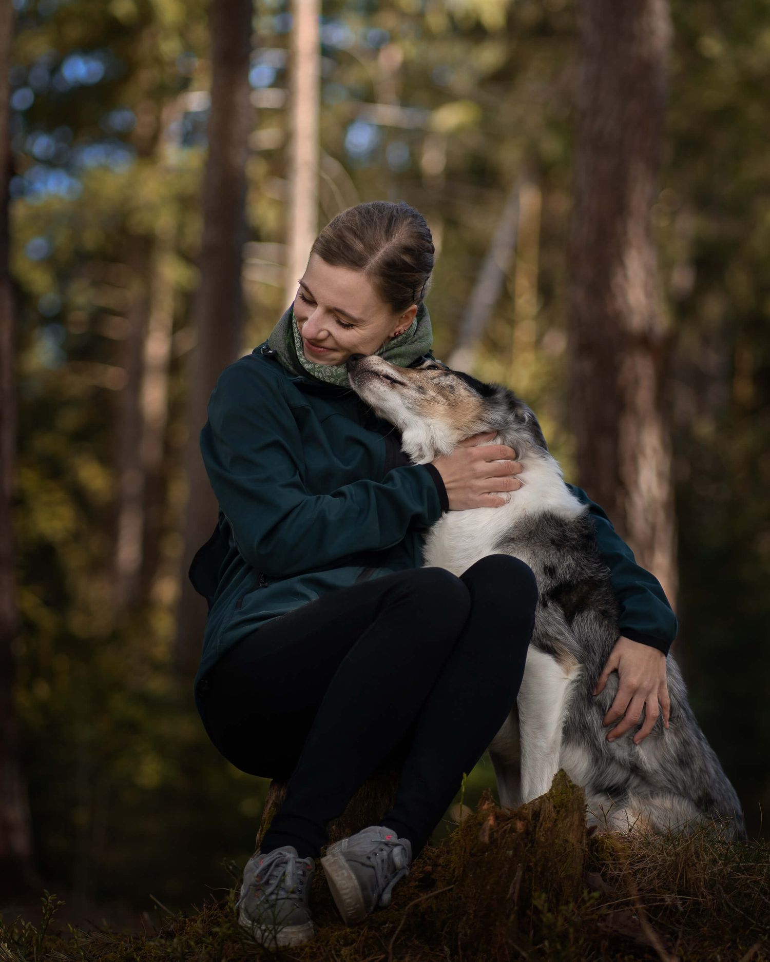 A smiling woman in a dark teal jacket hugs her blue merle Border Collie dog in a sunlit forest.