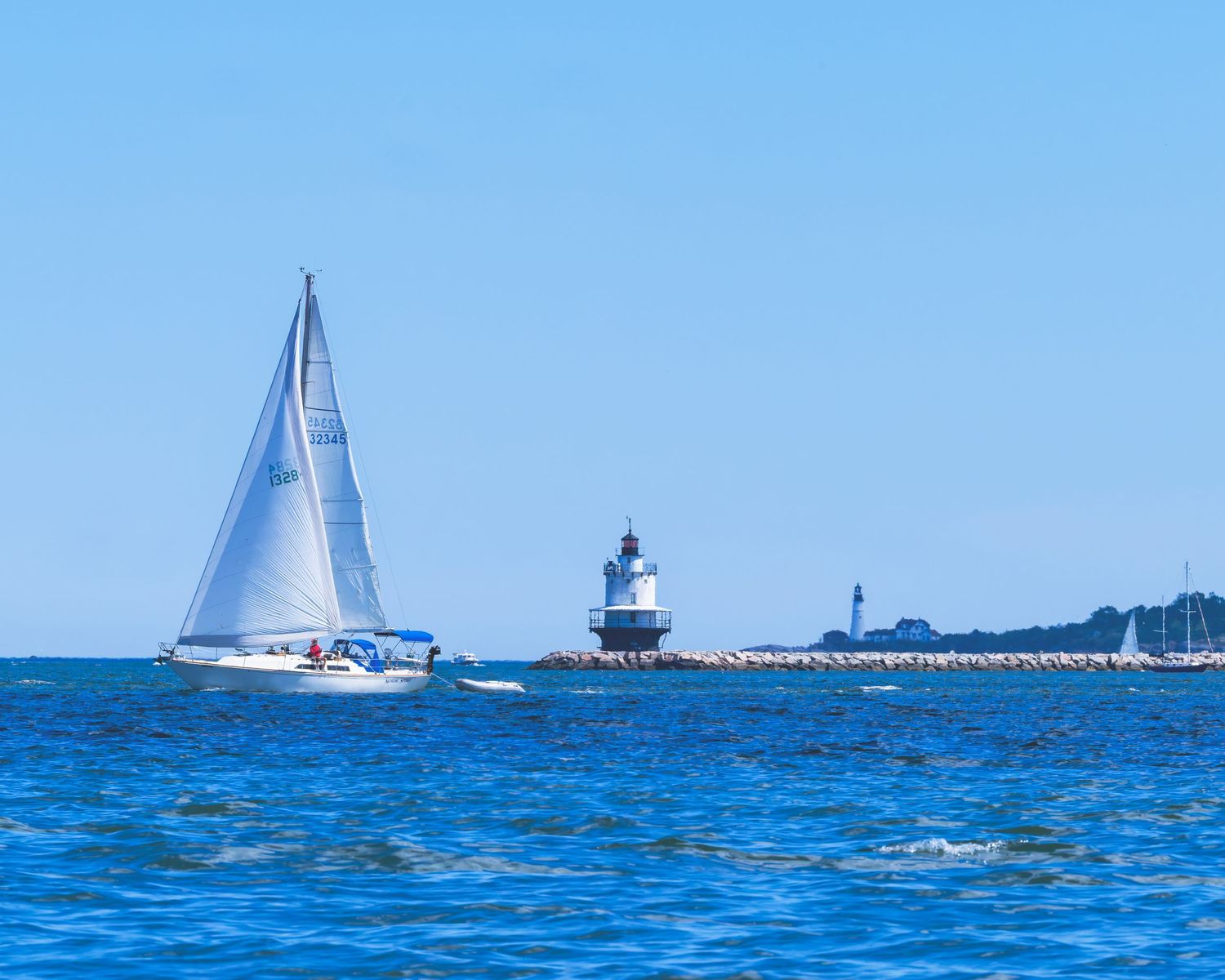 White sailboat gliding past Spring Point Ledge lighthouse in portland harbor Maine on calm blue ocean waters under clear sky.