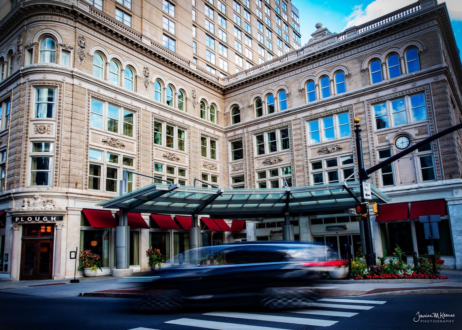 Blurred car passing a grand historic hotel building with green awnings and red accents on a busy city street.