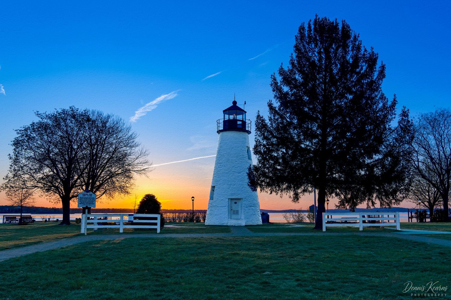 Havre de grace light house at sunrise with silhouetted trees and a colorful blue and orange sky reflecting on calm water.