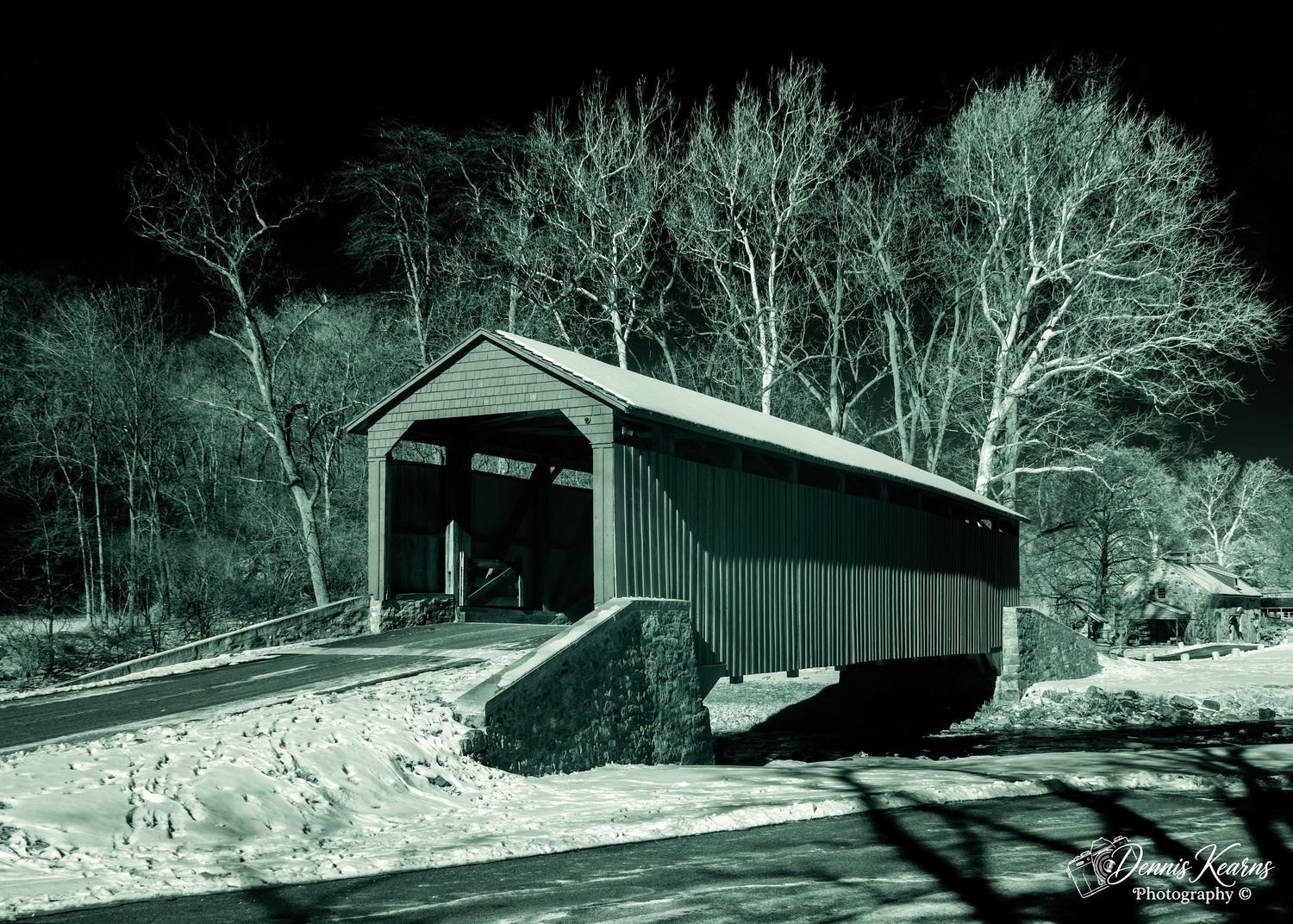 A vintage covered wooden bridge dusted with snow, surrounded by bare winter trees in a moody teal-toned night photograph.