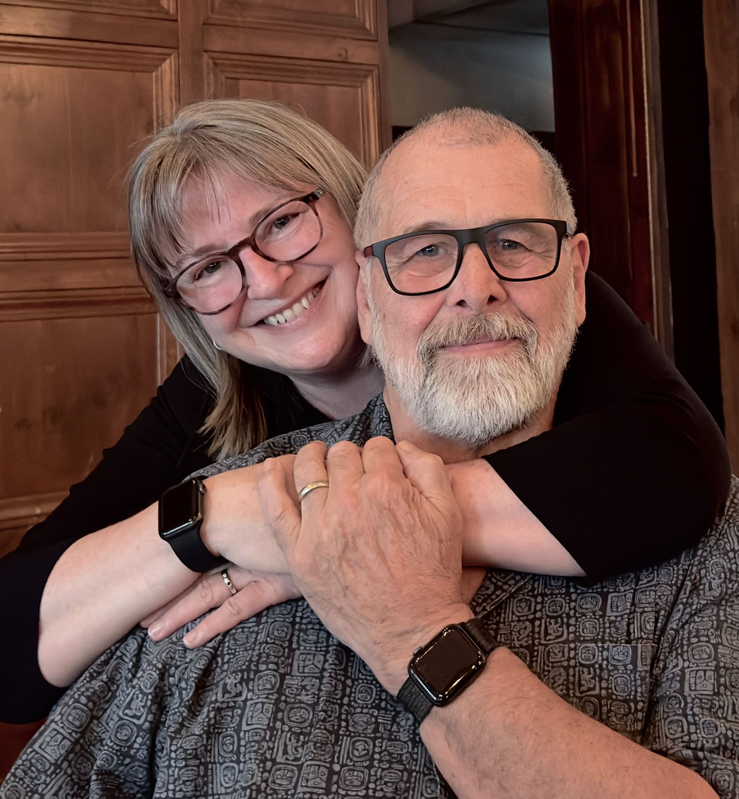 Happy loving couple wearing glasses smiling and warmly embracing each other.  The woman is hugging the man from behind affectionately.