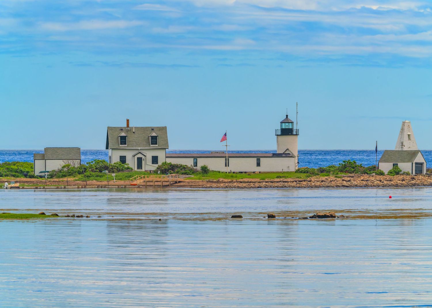 Goat Island Lighthouse complex with keeper's house, American flag, and white beacon reflected in calm harbor waters.