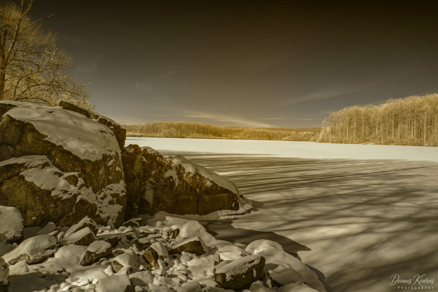 Infrared image of a snow-covered frozen lake with rocky shoreline and golden trees in a winter landscape.