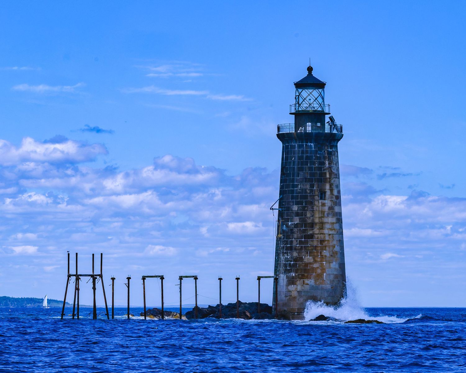 Ram Island Ledge lighthouse Stone tower rising from rocky shore with pier remnants and crashing waves under blue sky.