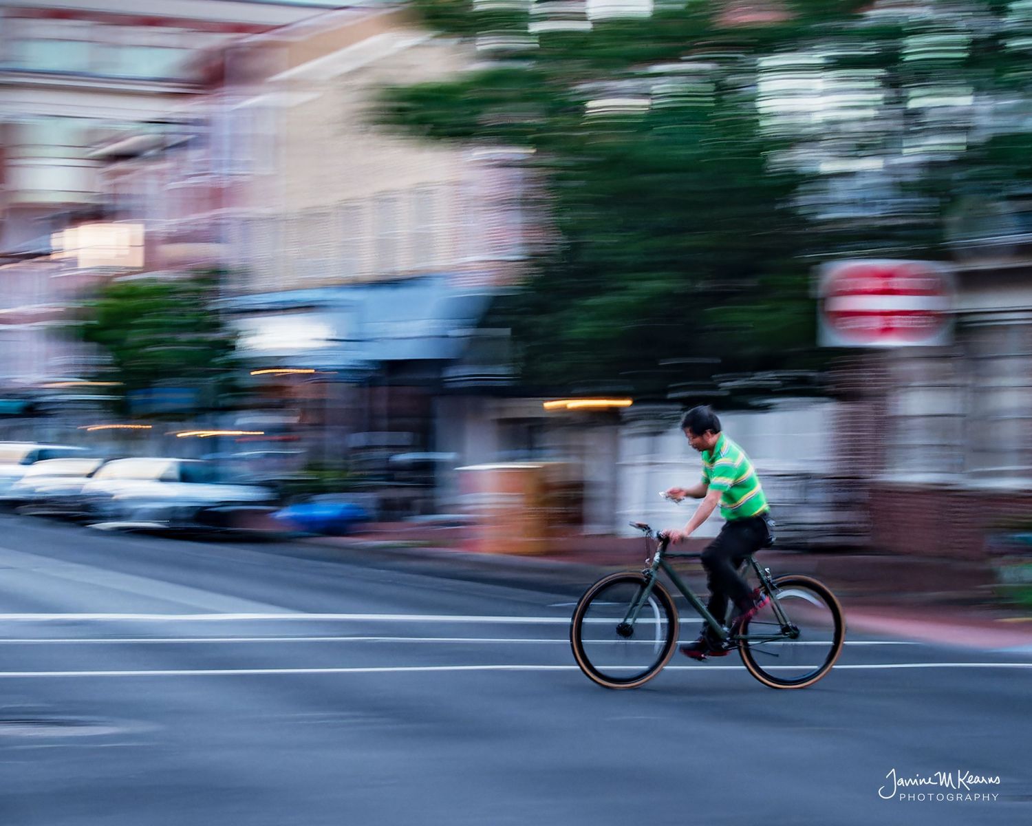 Cyclist in green shirt rides bike down urban street, captured with motion blur panning technique at dusk.