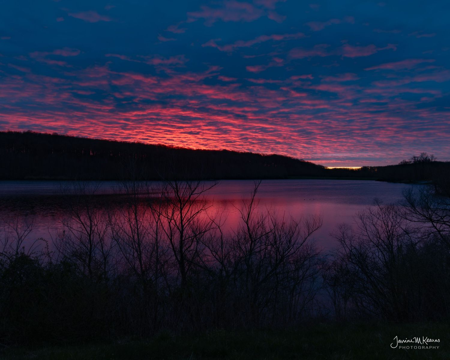 Vivid red and pink sunset over a calm lake with silhouetted trees and dramatic cloud formations in twilight sky.