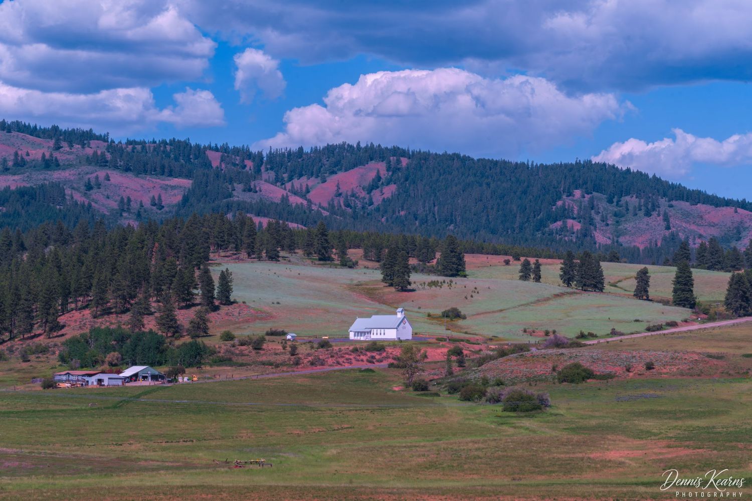 a view of the valley with Swauk-Teanaway Grange Cle Elum, WA nestled among evergreen trees and rolling hills under dramatic cloudy skies.