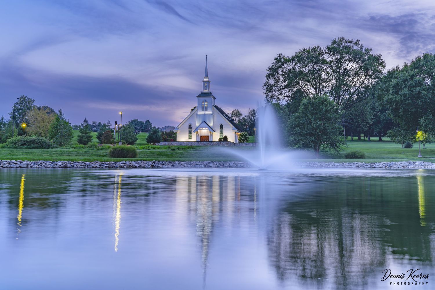 long exposure of the willow chapel taken at dusk, with reflections a fountain,  lush green trees and  a dramatic surreal sky.