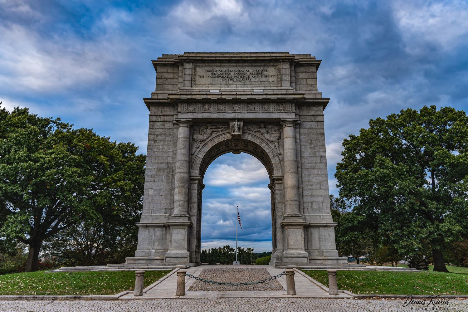 Historic stone arch monument stands against dramatic cloudy sky, framed by lush green trees at Valley Forge Park.