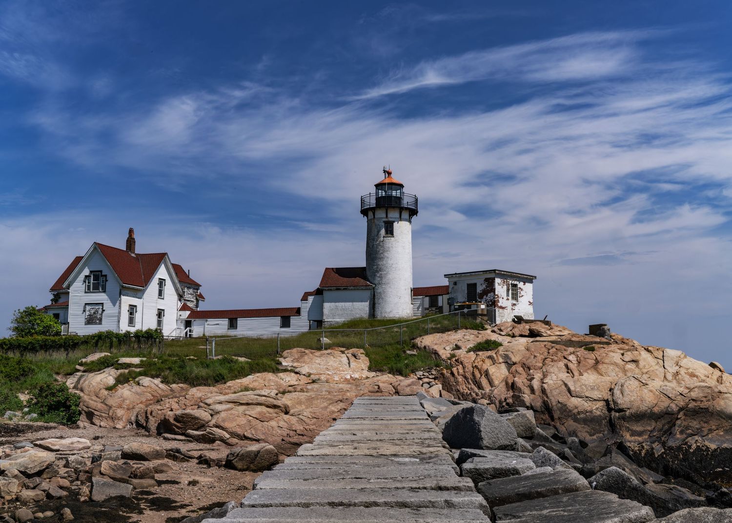 Gloucester Eastern Point Lighthouse with its white tower and the  keeper's house on rocky New England coastline under dramatic blue sky.
