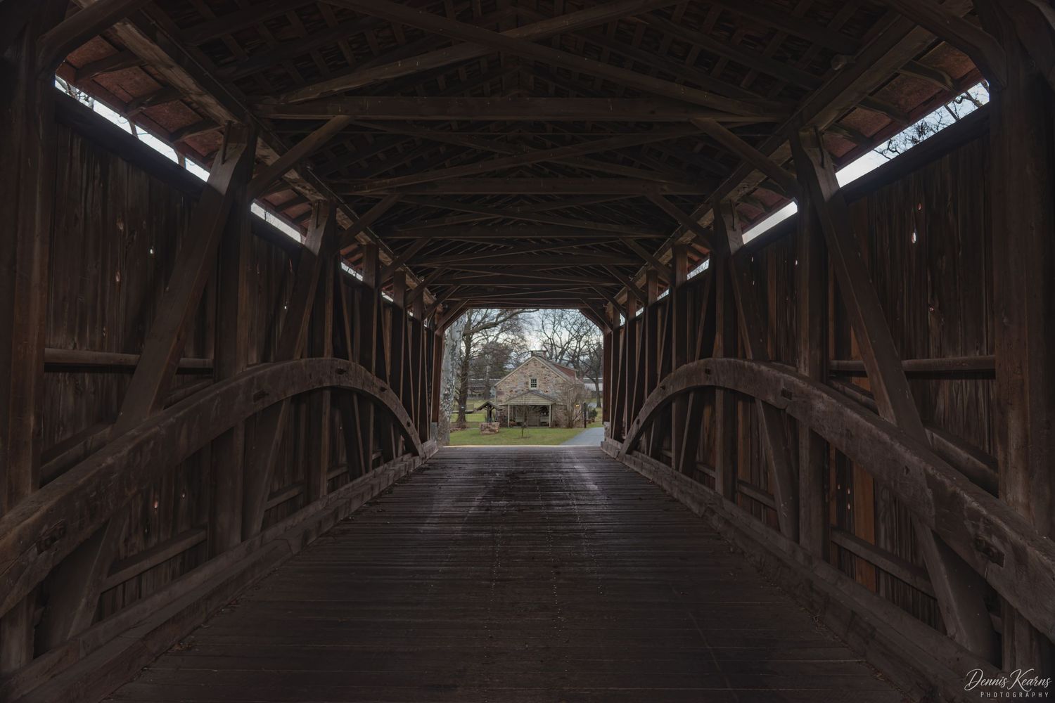 Interior view of a historic wooden covered bridge with arched timber framing, leading to a sunlit rural landscape.