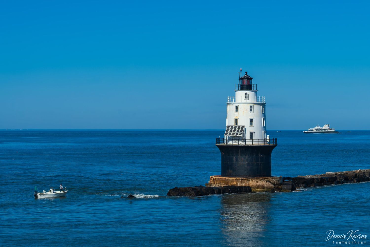 Harbor of Refuge Lighthouse on rocky pier surrounded by blue ocean waters with boats and a ship visible in the background.