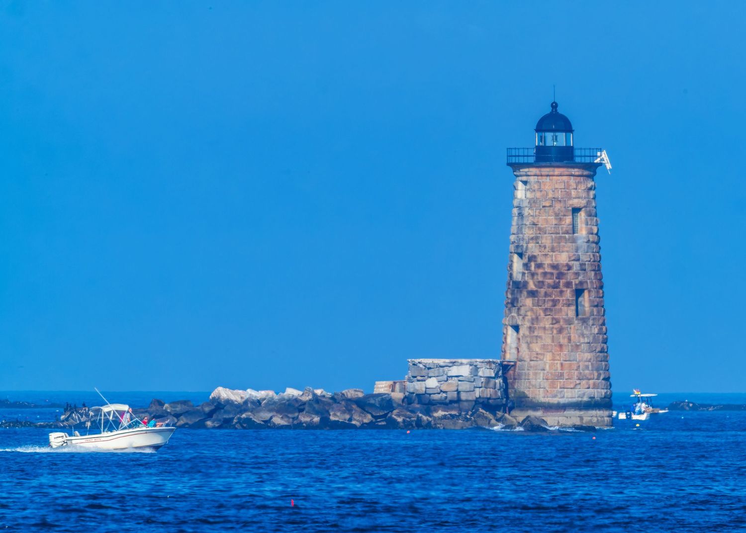 Historic granite Whaleback Lighthouse on small rocky island with passing tour boat on bright blue Maine ocean.