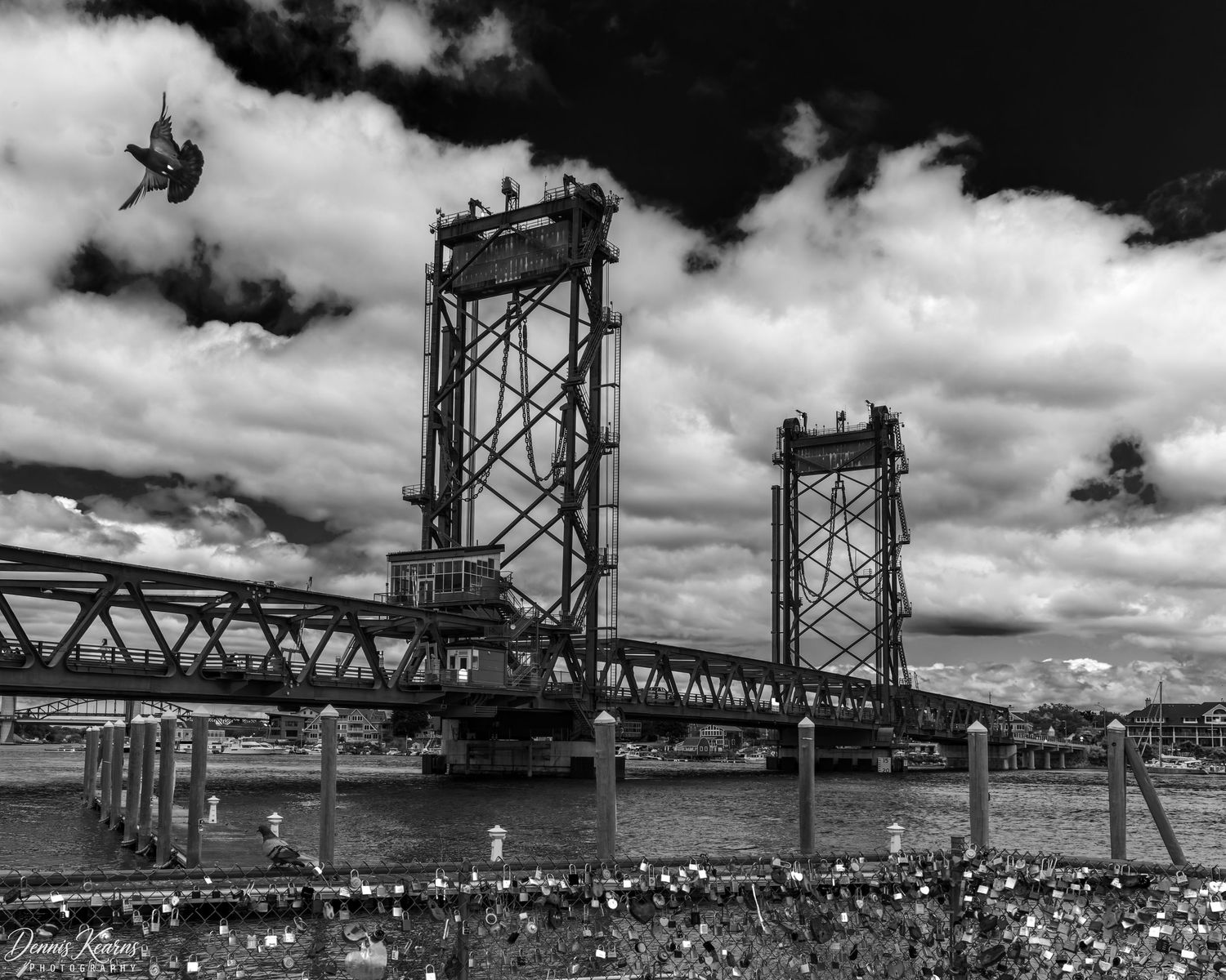 Black and white photo of a vertical lift bridge over a river with dramatic clouds and a bird in flight overhead.