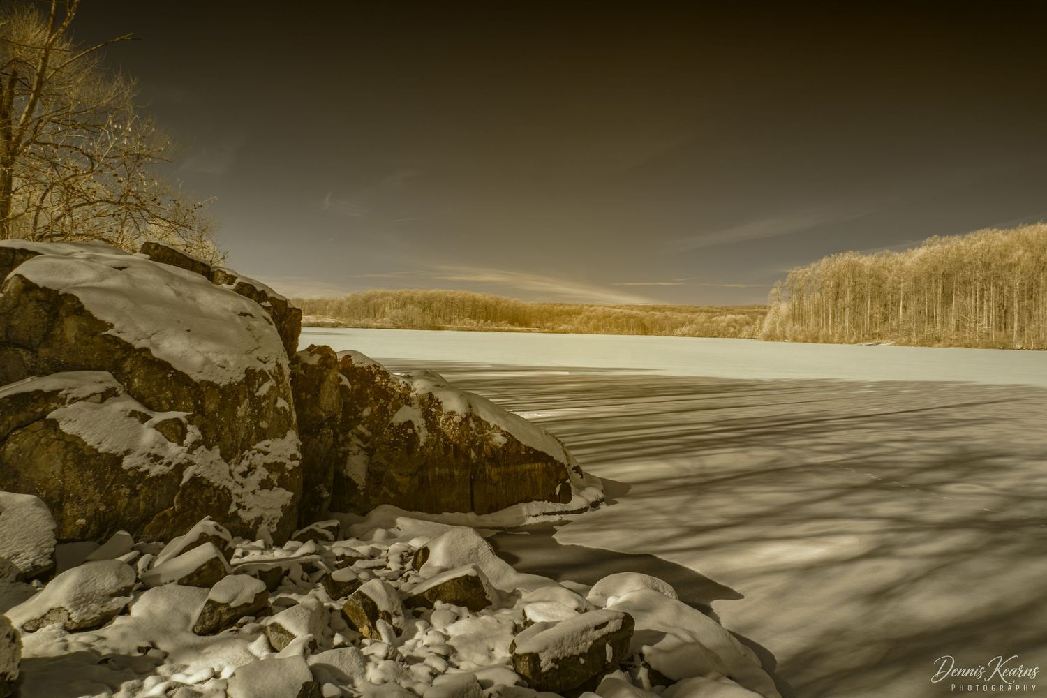 Infrared image of a snow-covered frozen lake with rocky shoreline and golden trees in a winter landscape.