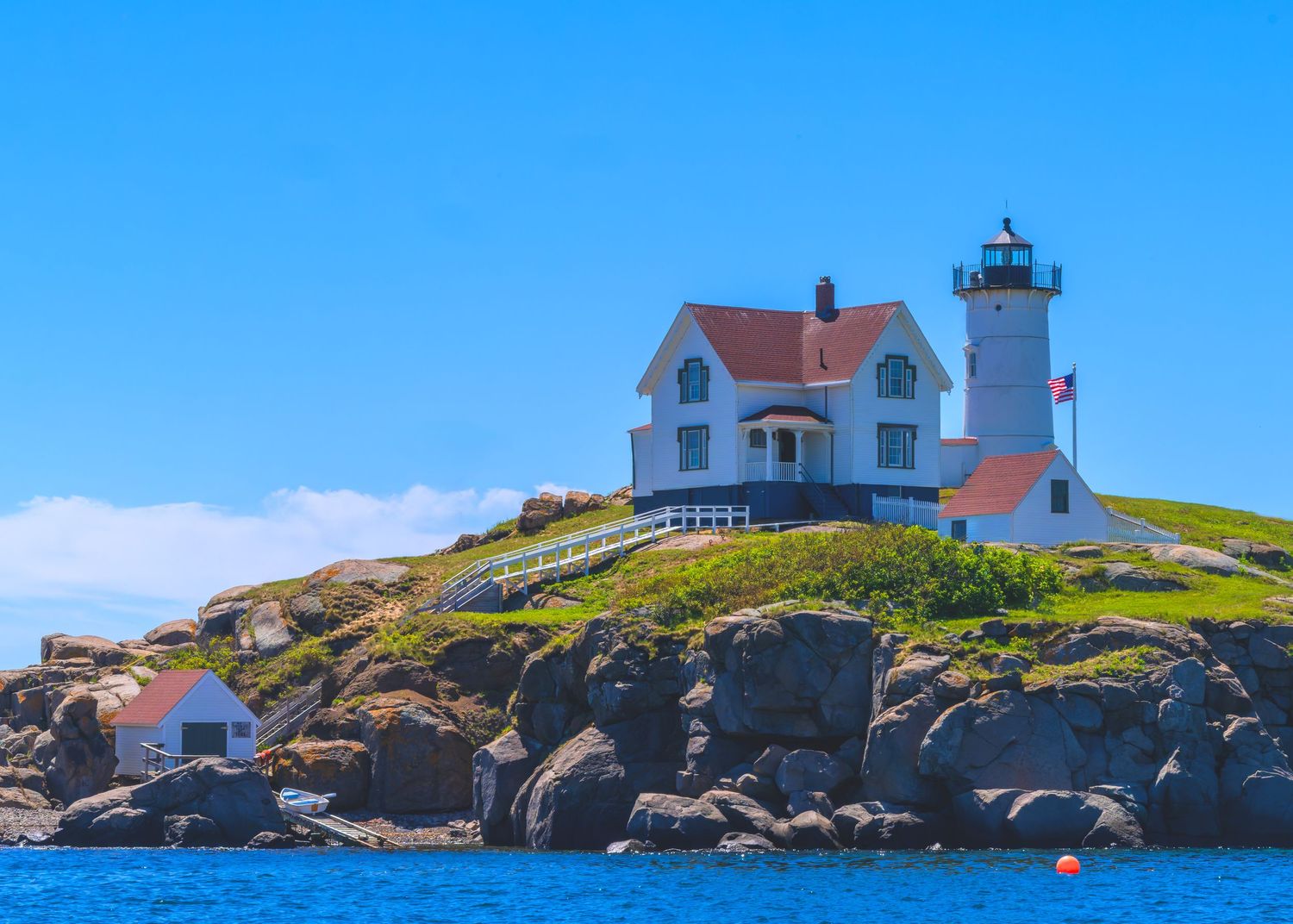 Nubble Lighthouse with white keeper's house perched on rocky coastal island under clear blue summer sky in Maine.