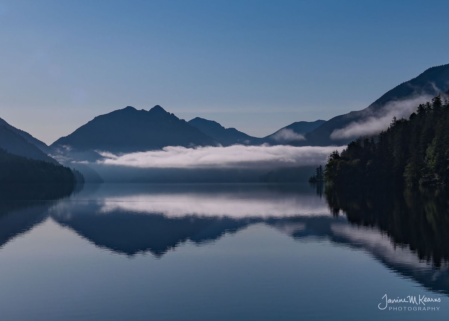 Lake Crescent with fog rolling over calm water surrounded by dark forested peaks under clear blue sky. Olympic Penninsula Washington
