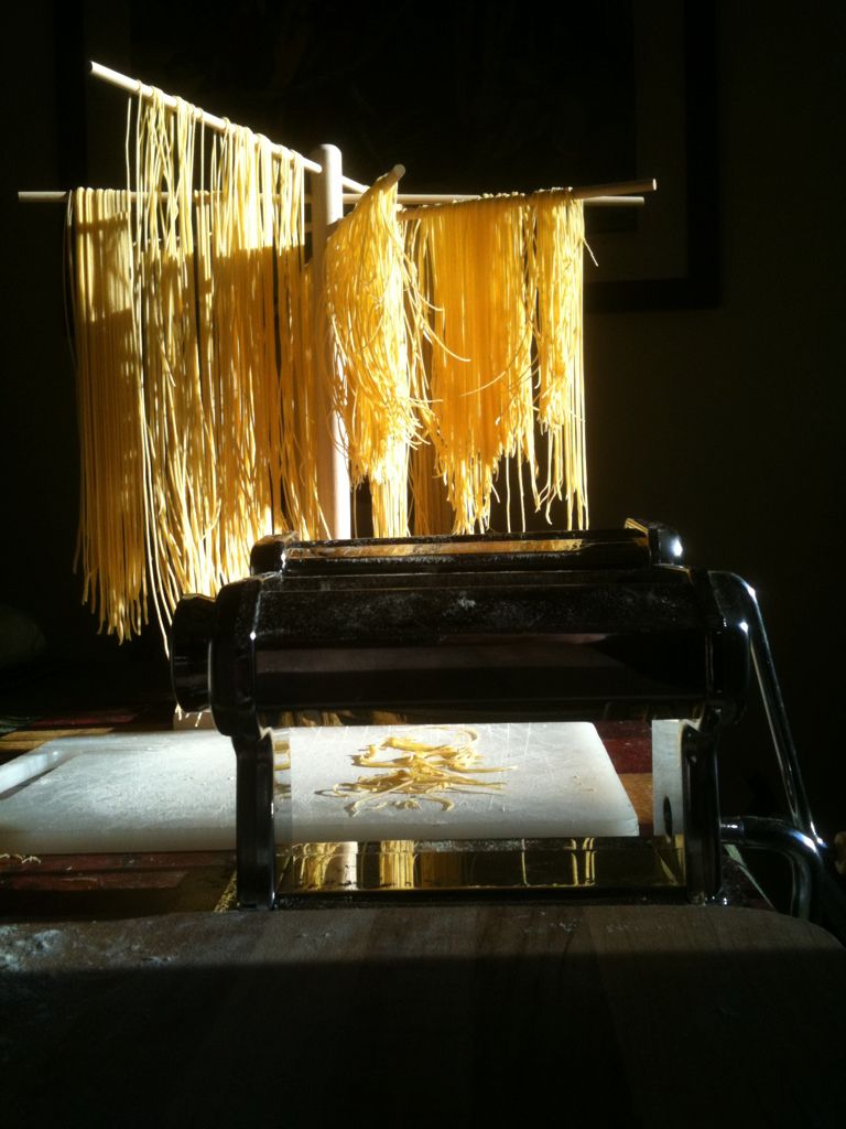 Fresh homemade pasta drying on a rack beside a vintage pasta machine on a floured surface in warm sunlight.