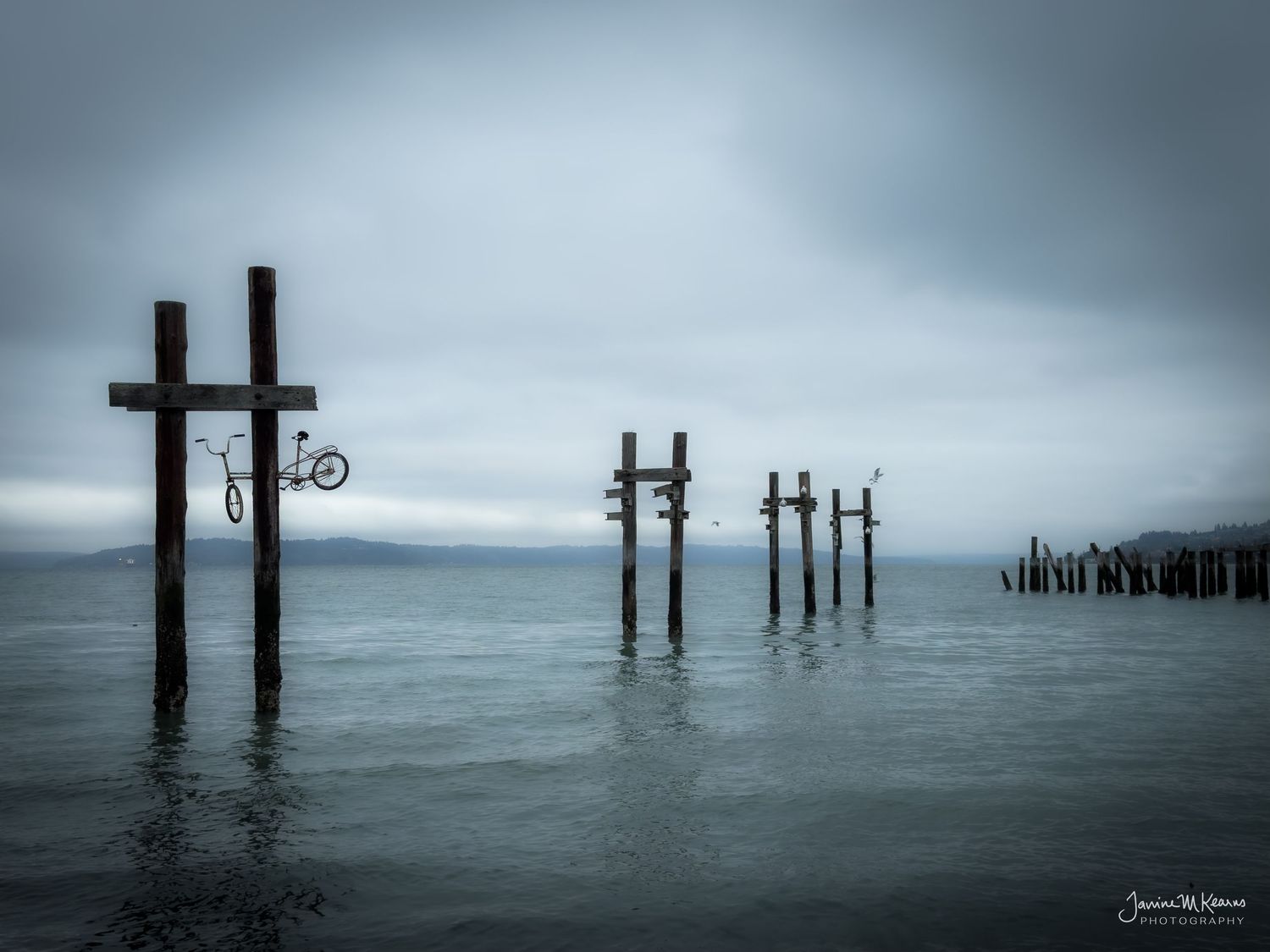 Abandoned pier pilings in calm gray water with a bicycle mounted on a cross under overcast skies.