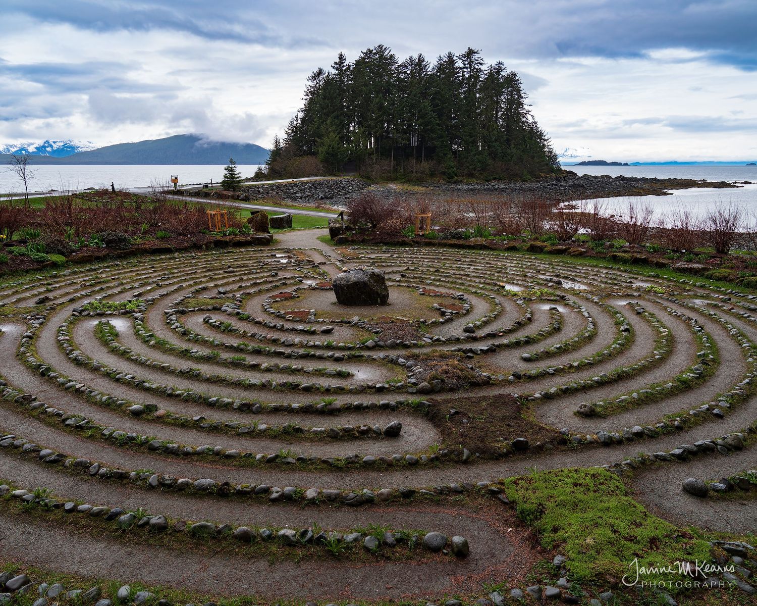 Stone labyrinth garden by the ocean with evergreen island backdrop under cloudy skies in Pacific Northwest.