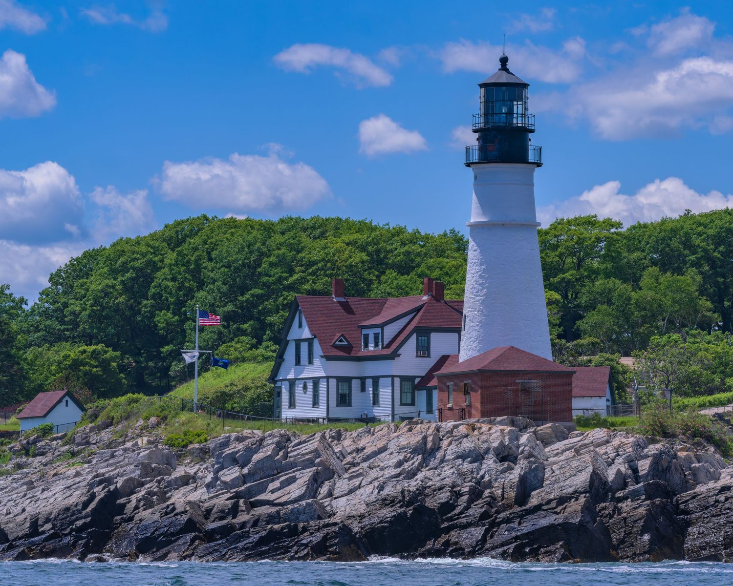 Portland Head Light historic lighthouse with red-roofed keeper's cottage on rocky Maine coast with lush green trees.