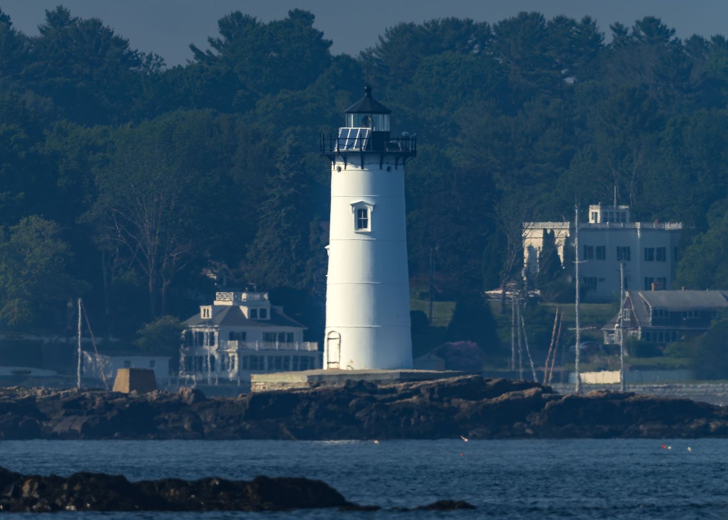 White colonial Portsmouth Harbor lighthouse and keeper's quarters on rocky New Hampshire coastline surrounded by lush green trees.