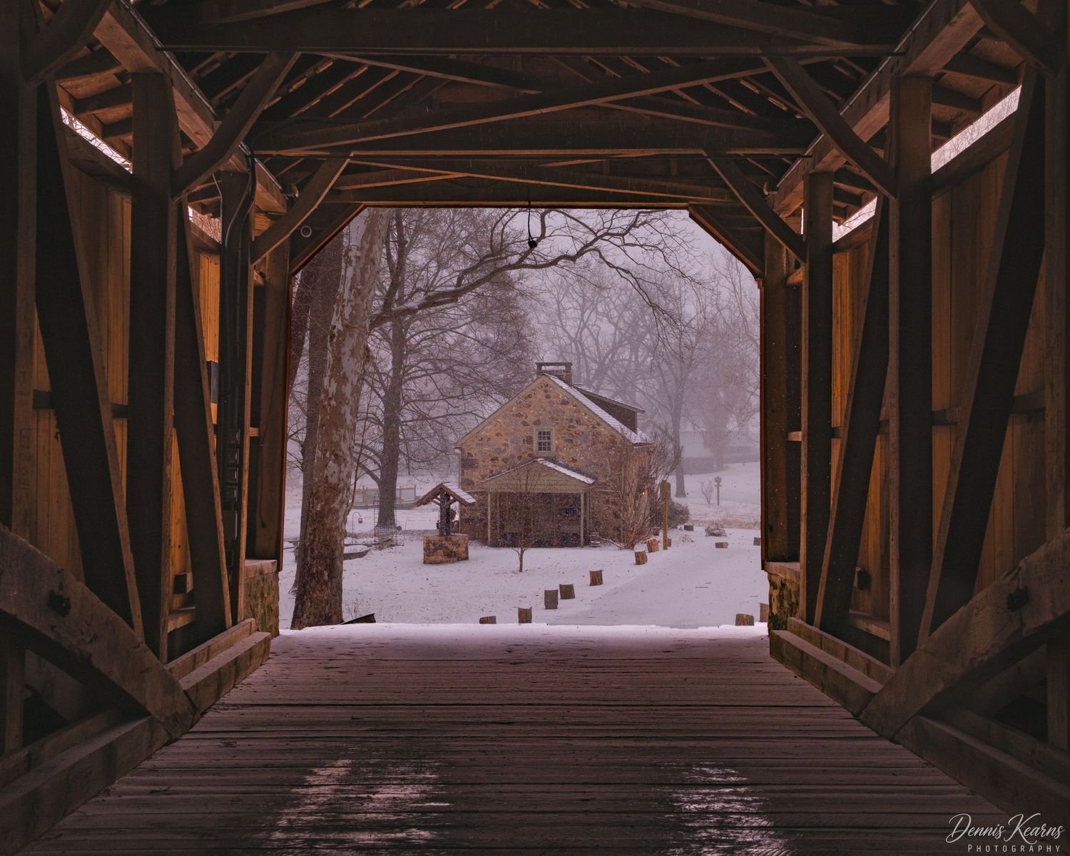 Snow-covered covered wooden bridge framing a historic stone building in a serene winter landscape.
