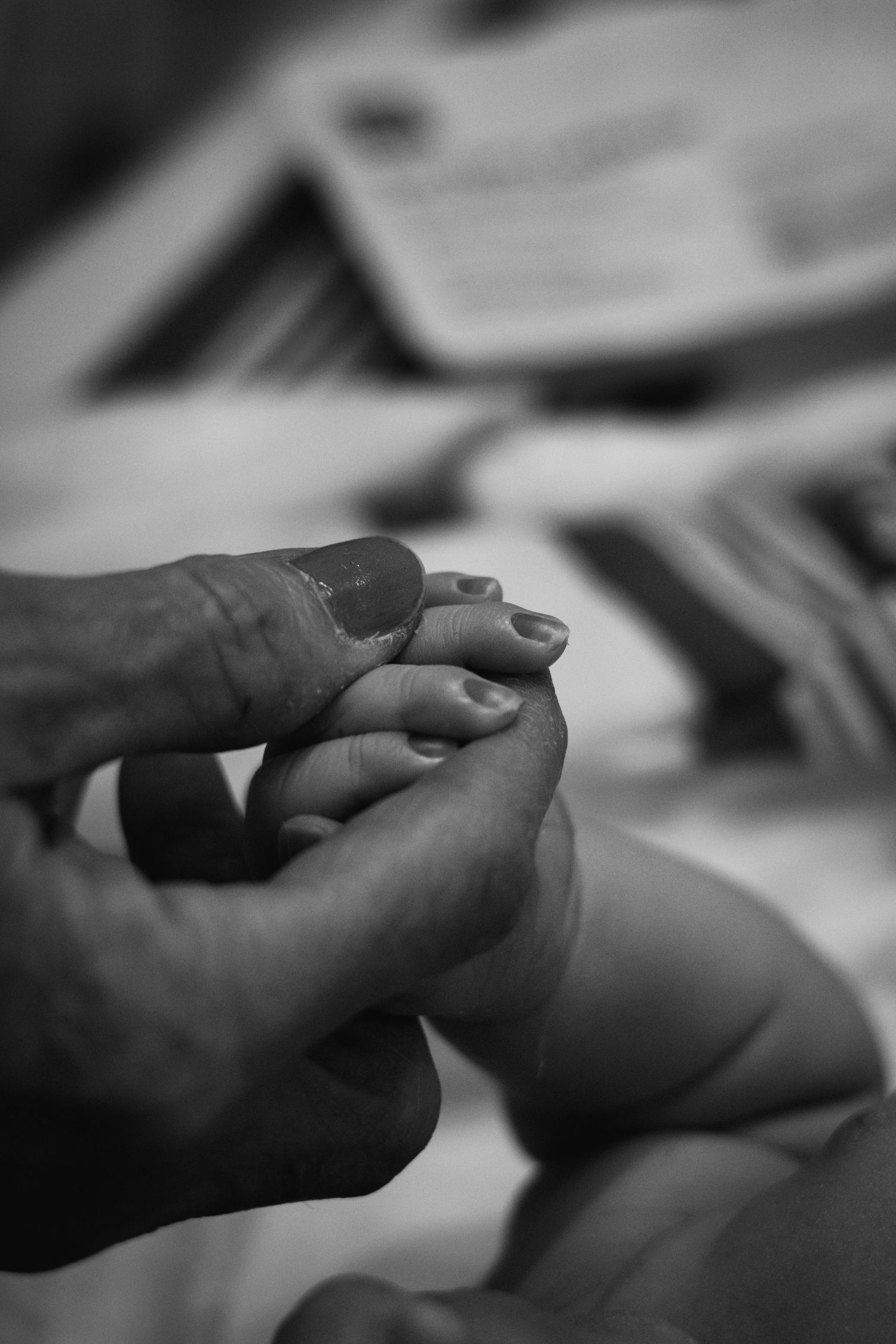 Black and white close-up of adult hand gently holding tiny newborn baby foot in hospital setting.