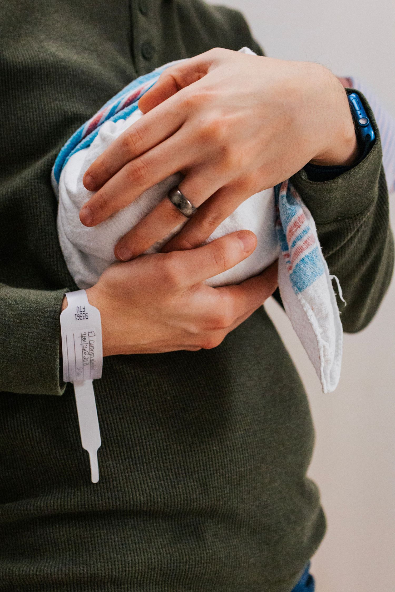 Close-up of parent's hands with wedding ring and hospital bracelet cradling swaddled newborn baby.