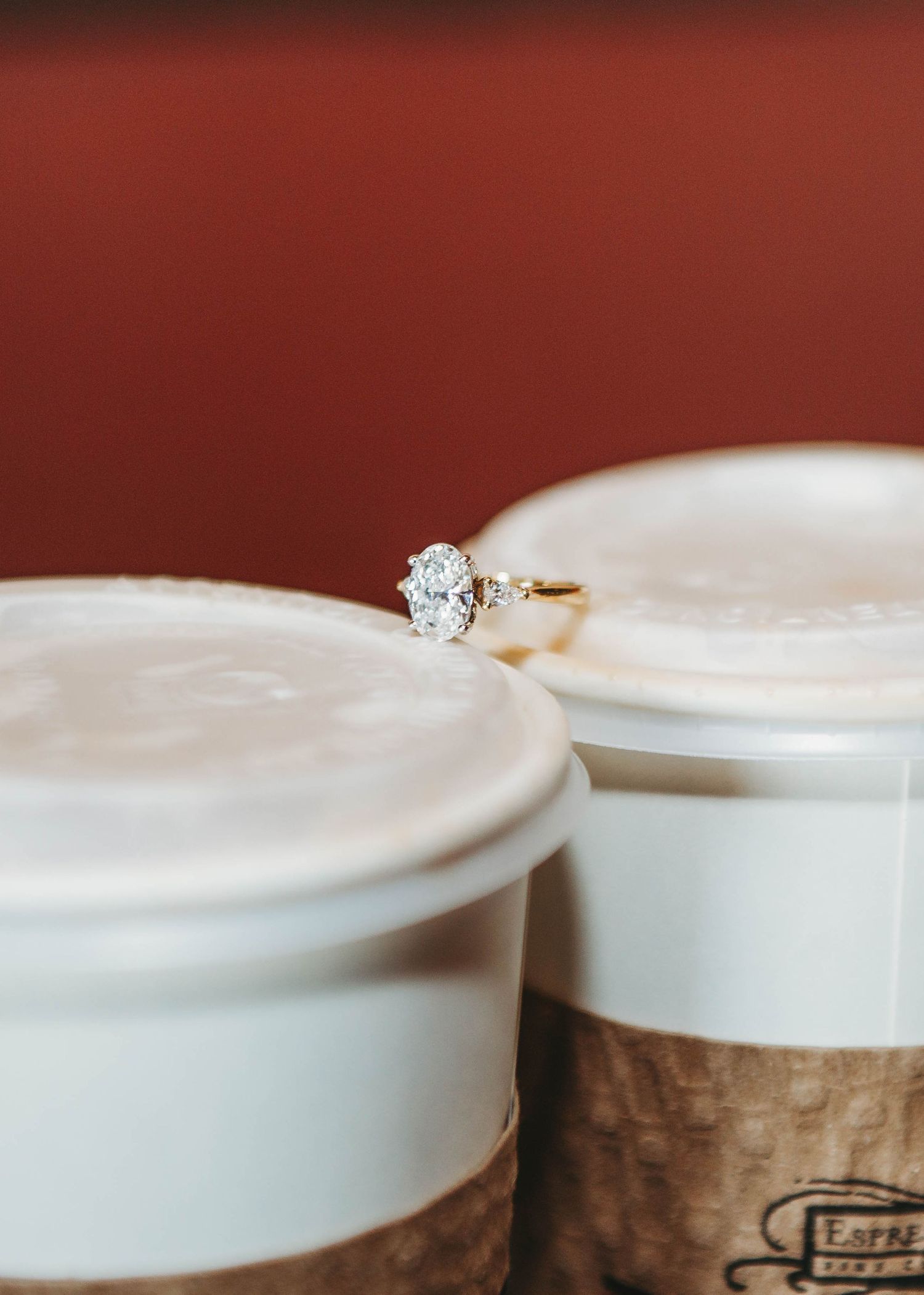 Close-up of an oval diamond engagement ring balanced between two coffee cups on a red background.