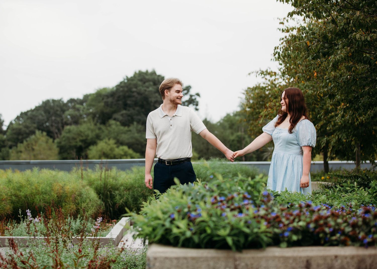 Couple holding hands in lush garden park on overcast day during engagement session.