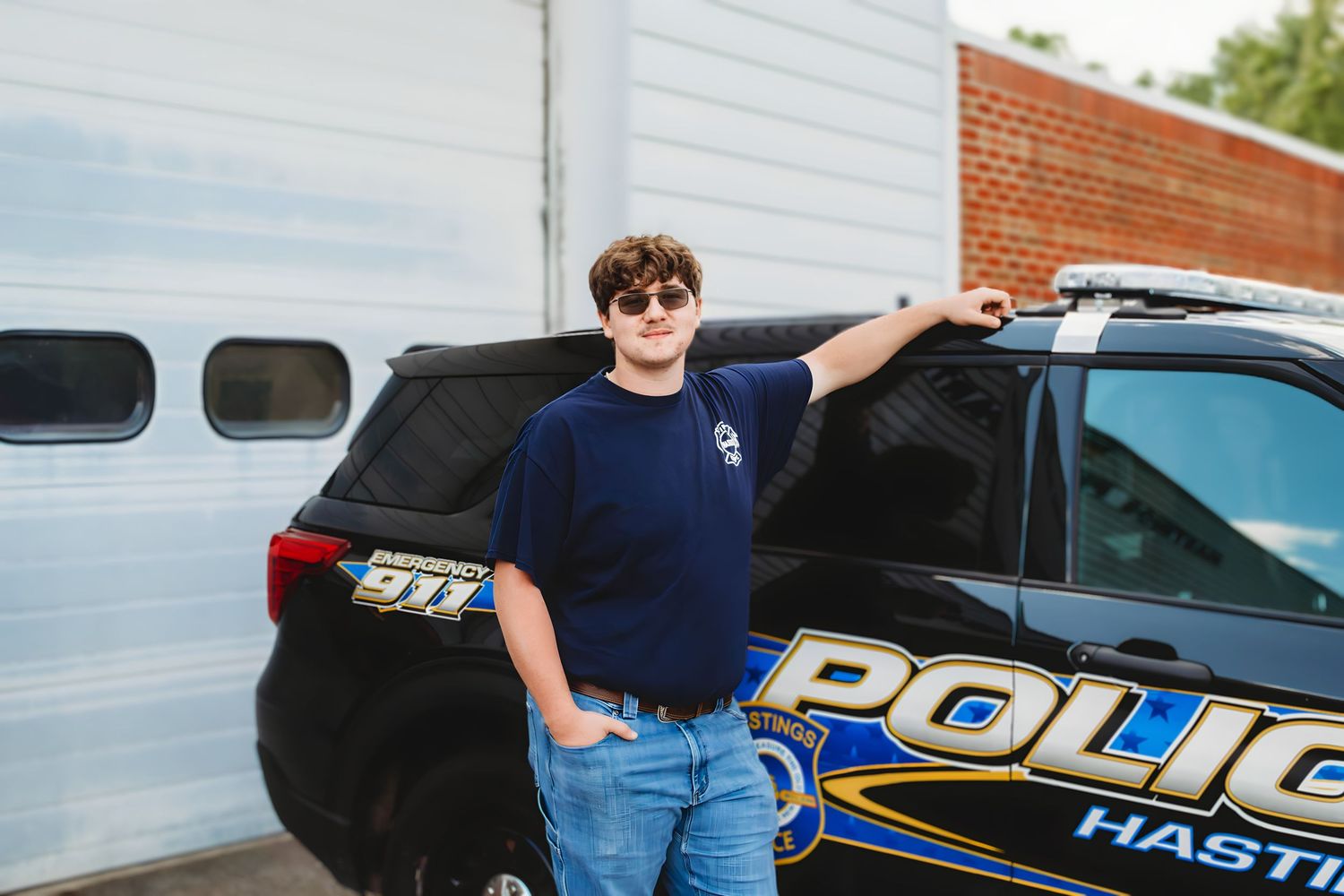Young man in navy police shirt leaning against a Hastings police SUV near a garage.