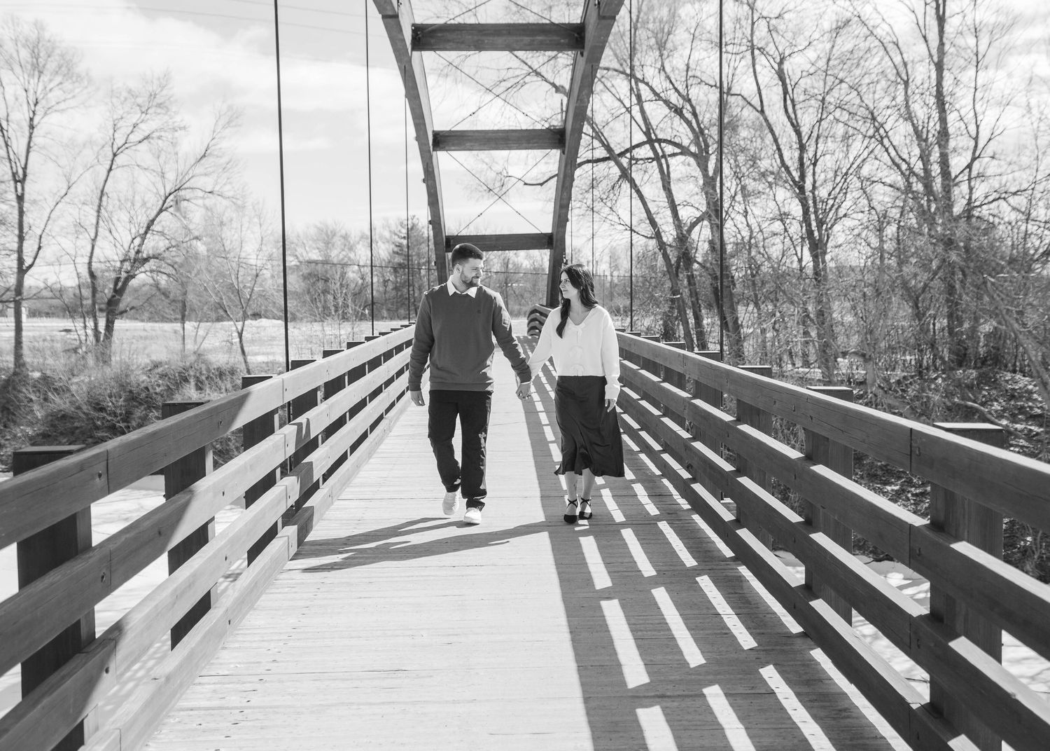 Black and white photo of couple walking hand in hand across a wooden bridge in winter.