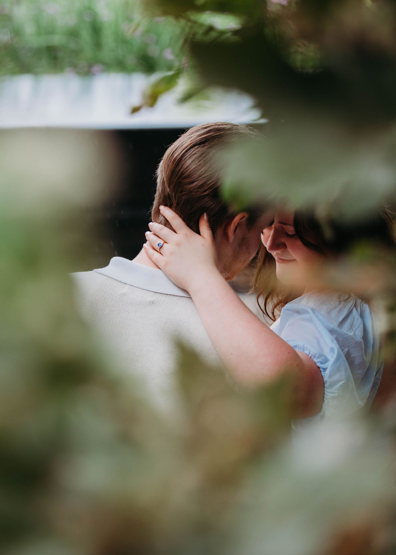 Romantic couple sharing an intimate moment framed by soft green bokeh leaves outdoors.