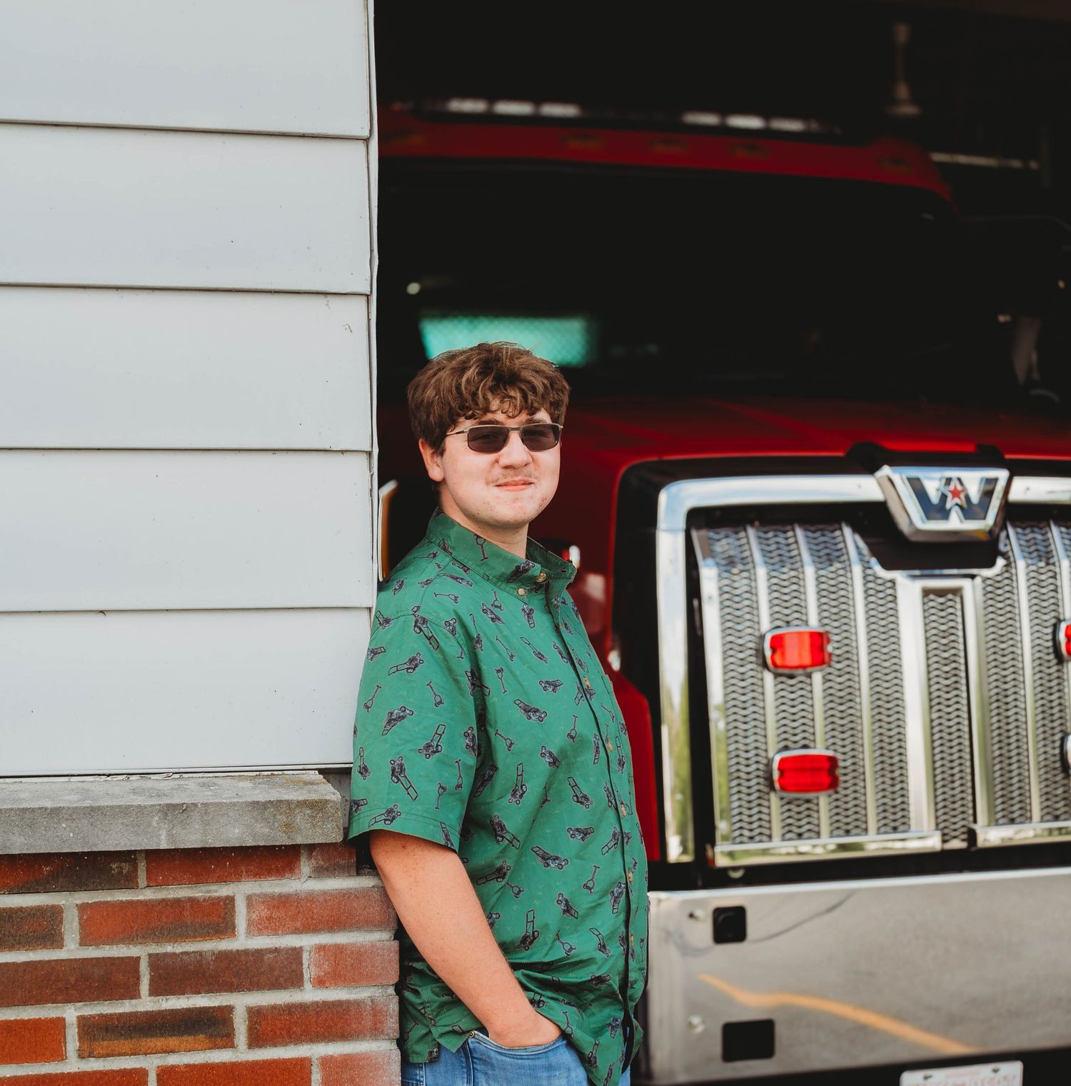 Smiling teen in green printed shirt leaning against a red Western Star truck by a brick building.