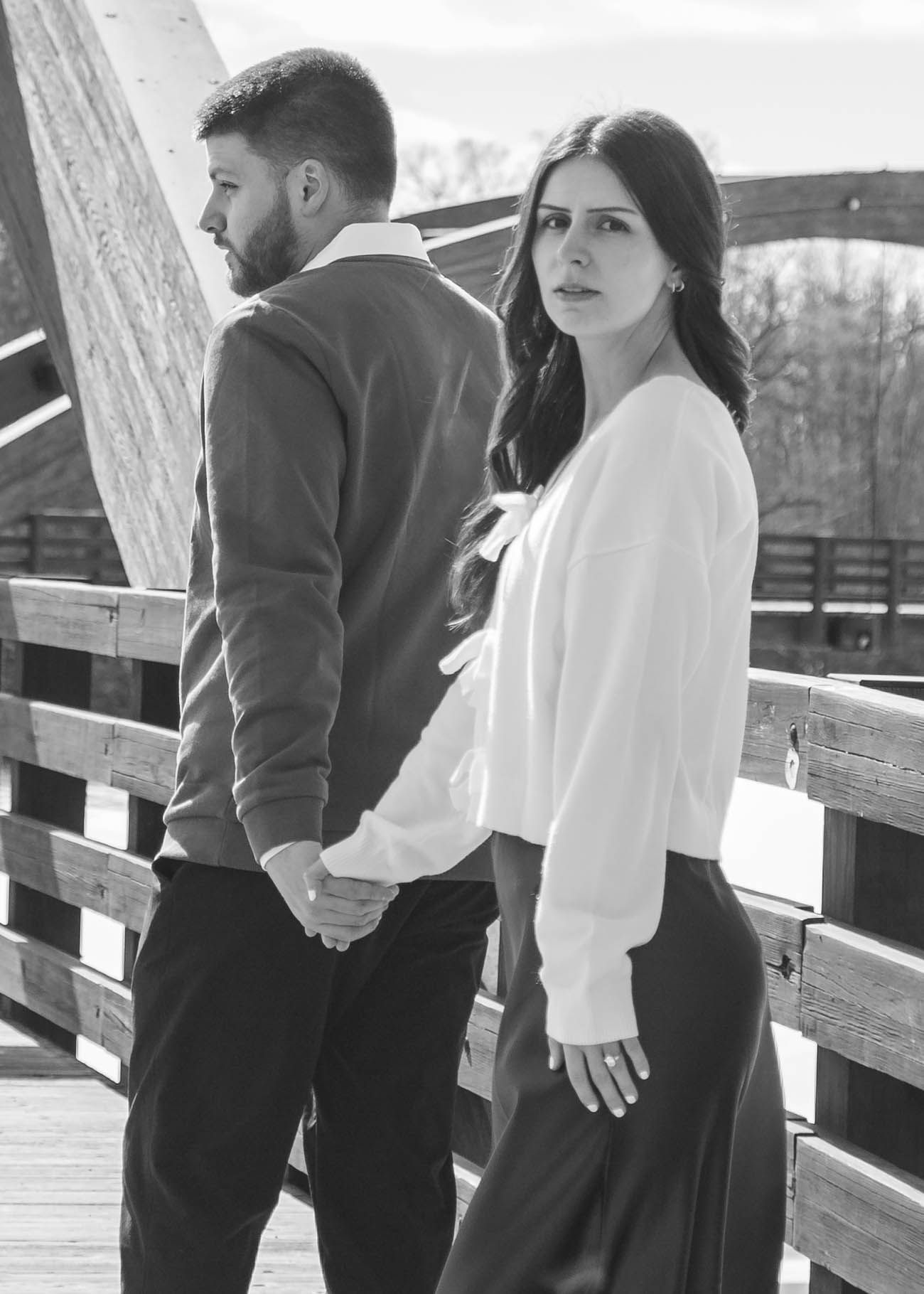 Black and white photo of a couple holding hands on a wooden bridge, looking back.