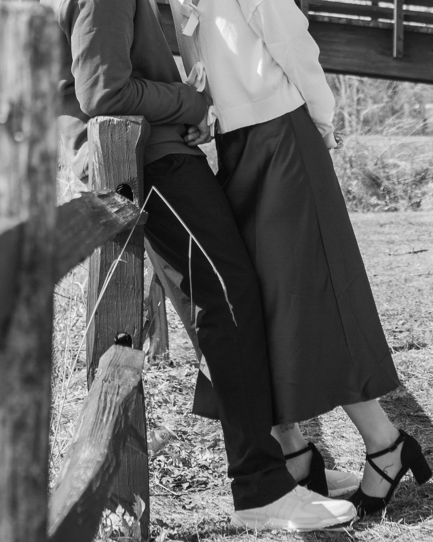 Black and white close-up of couple leaning against a wooden fence, woman wearing black heels.