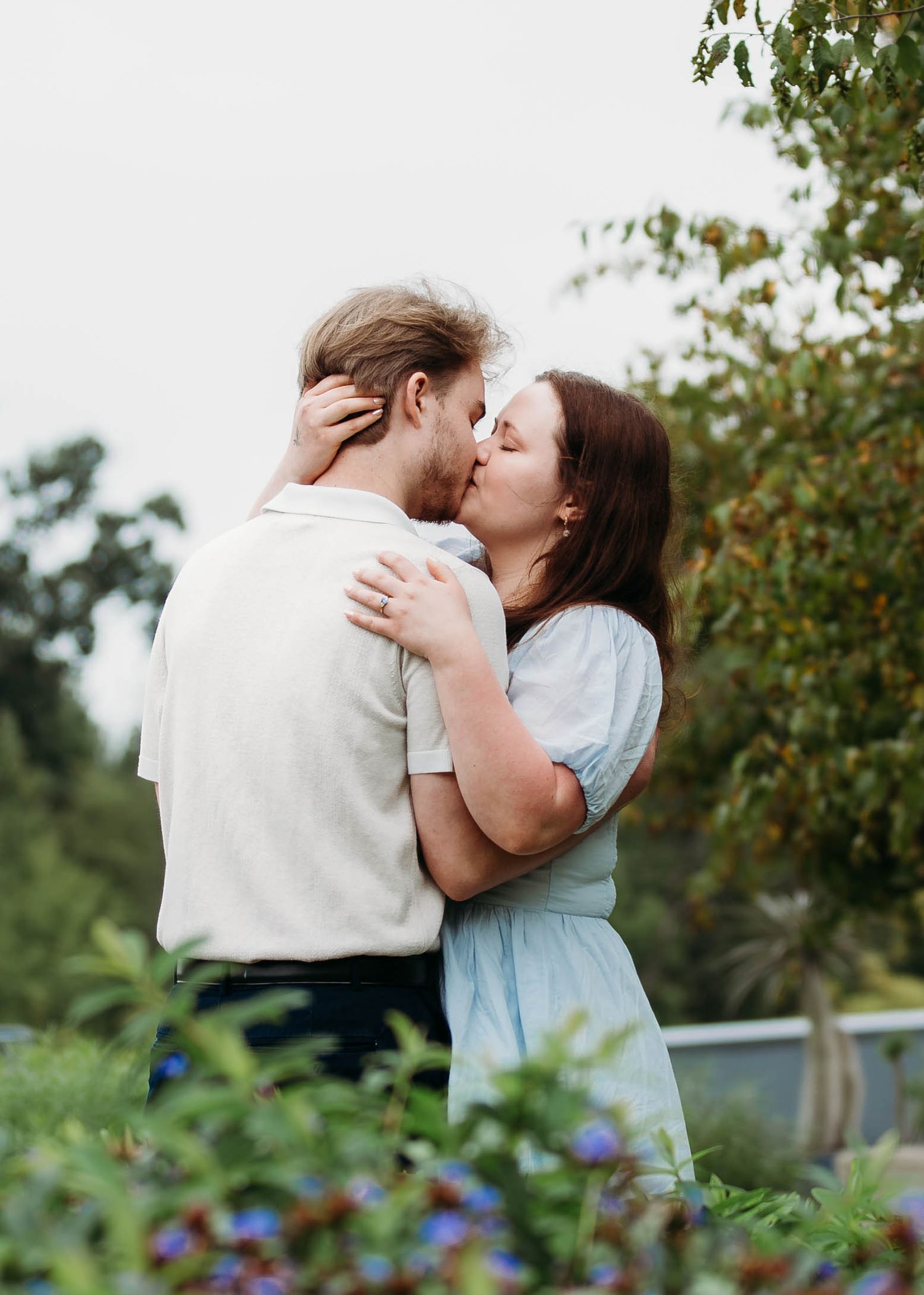 Romantic couple sharing a kiss outdoors surrounded by green trees and purple flowers.