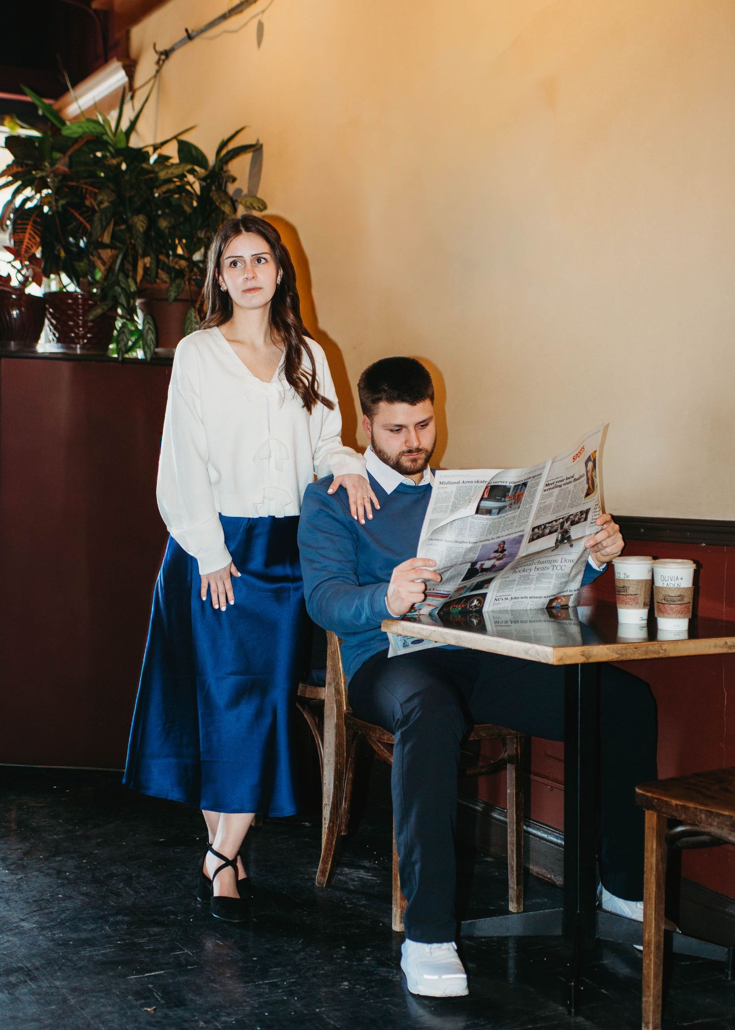 Woman in blue satin skirt stands beside seated man reading newspaper in cozy cafe.