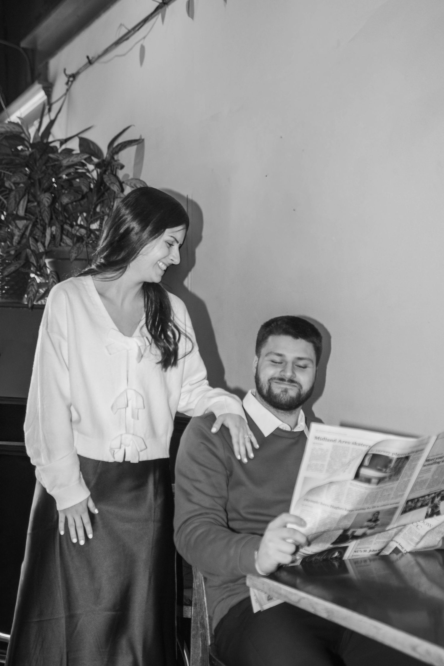 Black and white photo of couple in cafe, woman smiling as man reads newspaper.
